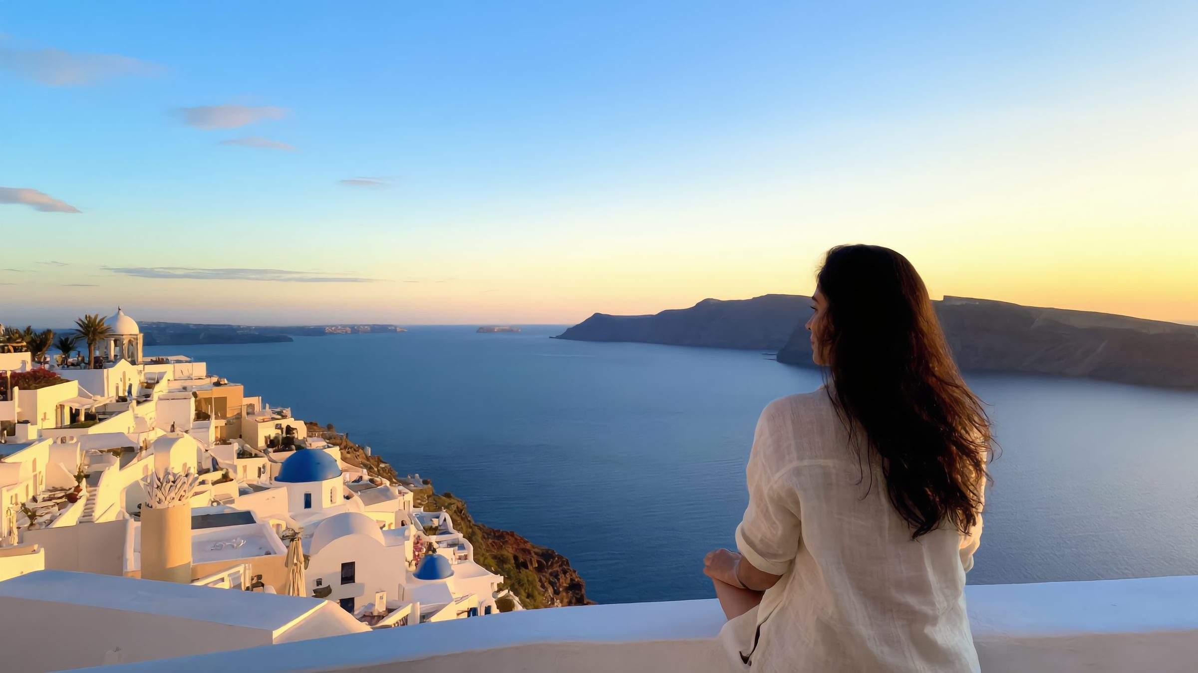 Lia sits on a whitewashed wall above the caldera in Oia at golden hour, seen from behind, with the sea and the white houses below.