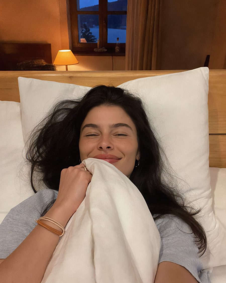 Lia wakes up smiling in a cozy wooden room in Hallstatt, with the lake in the window behind her.